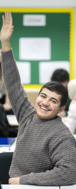 A male student in a gray sweater raises his hand enthusiastically in a classroom. He smiles while other students work in the background A male student in a gray sweater raises his hand enthusiastically in a classroom. He smiles while other students work in the background